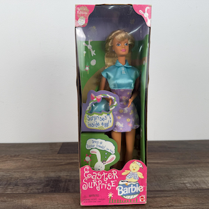 May include: A Barbie doll in a clear plastic box. The doll has blonde hair and wears a blue top and a purple skirt. The box is labeled "Easter Surprise Barbie" with a pink and green design.