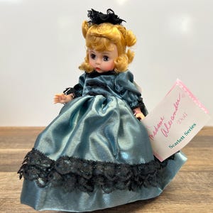 May include: A blue and black porcelain doll wearing a blue satin dress with black lace trim. The doll has blonde hair and blue eyes. The doll is labeled "Madame Alexander Doll Scarlett Series".