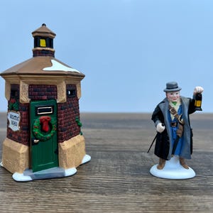 May include: A small brick building with a green door and a wreath, labeled "Welcome Home", and a figurine of a man in a blue coat and hat holding a lantern.
