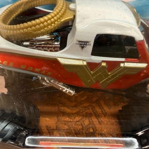 May include: A toy monster truck with a red, white, and gold Wonder Woman design. The truck features the Wonder Woman logo and the words "Wonder Woman" on the side. The truck has large black tyres and a gold-coloured roll cage.