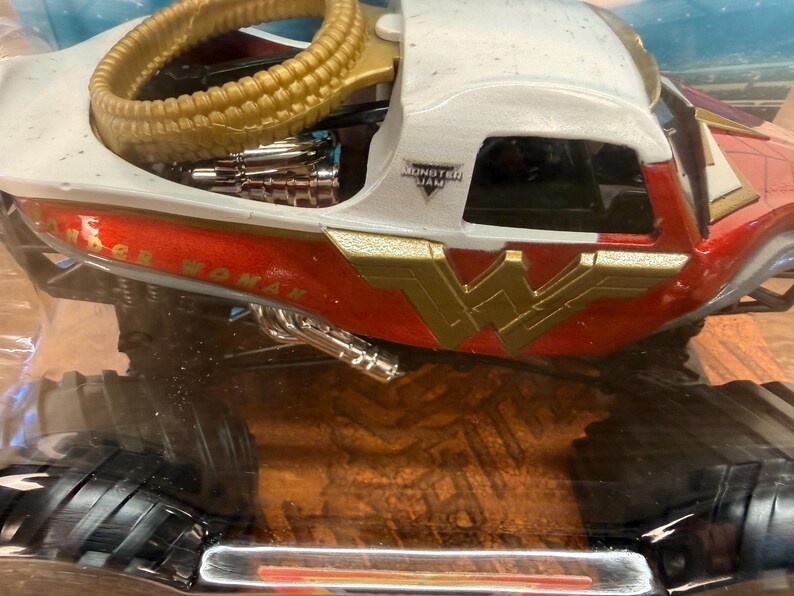 May include: A toy monster truck with a red, white, and gold Wonder Woman design. The truck features the Wonder Woman logo and the words "Wonder Woman" on the side. The truck has large black tyres and a gold-coloured roll cage.