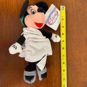 May include: A plush Mickey Mouse toy wearing a white toga and sandals. The toy is approximately 8 inches tall and has a green headband. The toy has a tag that reads "The Disney Store".