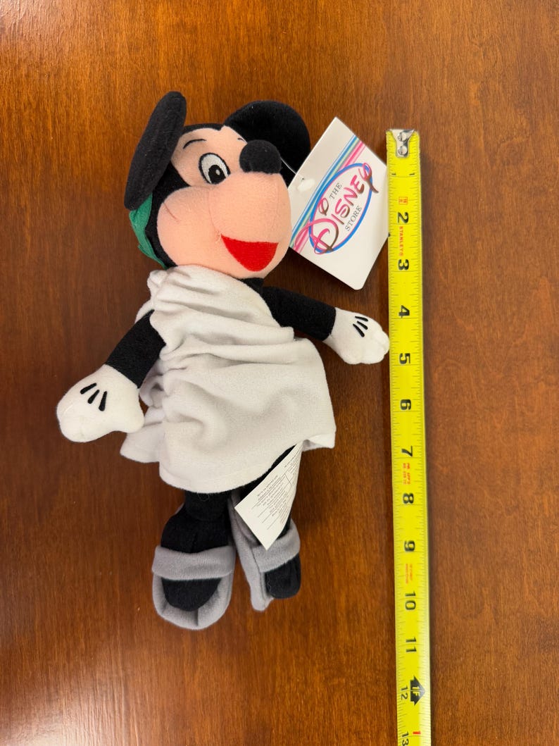 May include: A plush Mickey Mouse toy wearing a white toga and sandals. The toy is approximately 8 inches tall and has a green headband. The toy has a tag that reads "The Disney Store".