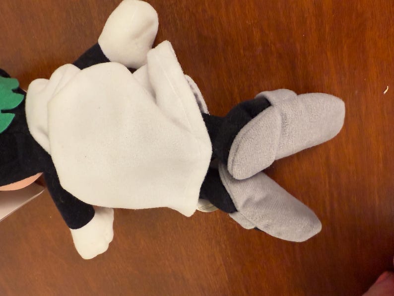 May include: A plush toy with a white body, black arms and legs, and a green head. The toy is wearing grey slippers.