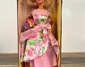 May include: A Barbie doll wearing a pink and white floral dress with a gingham pattern. She is holding a bouquet of flowers and has a pink bow in her hair. The doll is in a box with a pink hairbrush and a pair of pink shoes.