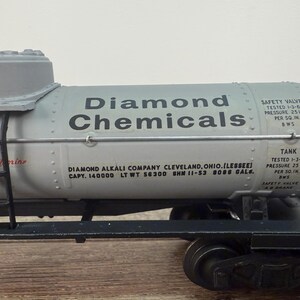 K&ouml;nnte beinhalten: Ein Modell-Eisenbahnwagen mit einem grauen Tank mit der Aufschrift "Diamond Chemicals". Der Tank hat schwarzen Text und eine Leiter. Der Wagen steht auf schwarzen R&auml;dern und Schienen. Der Text "Liquid Corine" ist sichtbar.