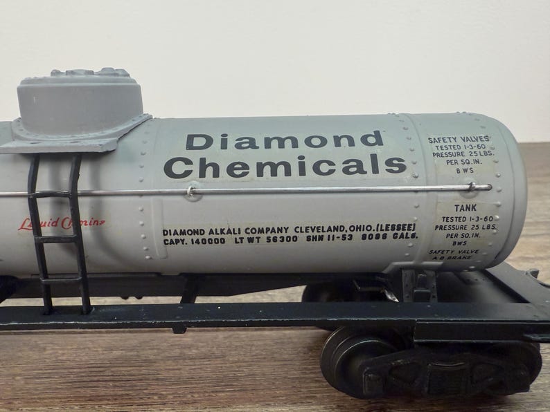 K&ouml;nnte beinhalten: Ein Modell-Eisenbahnwagen mit einem grauen Tank mit der Aufschrift "Diamond Chemicals". Der Tank hat schwarzen Text und eine Leiter. Der Wagen steht auf schwarzen R&auml;dern und Schienen. Der Text "Liquid Corine" ist sichtbar.