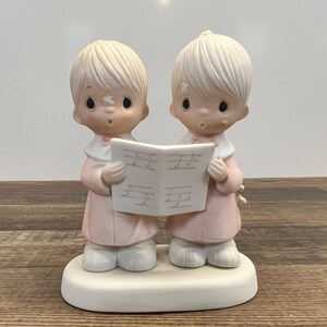 May include: A porcelain figurine of two children singing from a music book. They are wearing pink and white robes and have blonde hair. The figurine is on a white base.