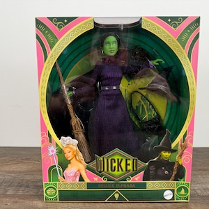May include: A boxed doll of Elphaba from the musical "Wicked." The doll has green skin, a purple dress, and a black hat. The box is pink and green with the word "Wicked" in a green font. A broom and a wand are also included.