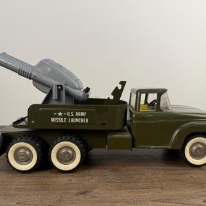 May include: Olive green toy truck with a missile launcher. The truck has white-walled tires and the text "U.S. ARMY MISSILE LAUNCHER" on the side. The missile launcher is gray.