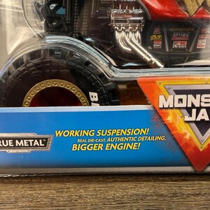 May include: A Monster Jam toy truck in a clear plastic package. The truck has a red and black body with a large tyre. The package has text that reads "WORKING SUSPENSION! REAL DIE-CAST, AUTHENTIC DETAILING, BIGGER ENGINE!" and the Monster Jam logo.