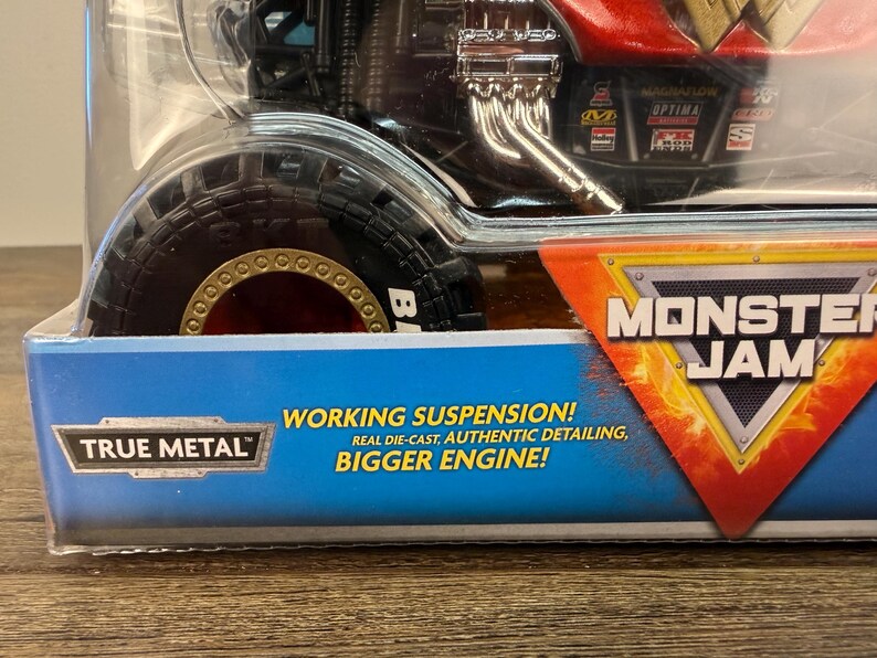 May include: A Monster Jam toy truck in a clear plastic package. The truck has a red and black body with a large tyre. The package has text that reads "WORKING SUSPENSION! REAL DIE-CAST, AUTHENTIC DETAILING, BIGGER ENGINE!" and the Monster Jam logo.
