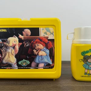 May include: A vintage yellow Cabbage Patch Kids lunchbox and thermos set. The lunchbox features a colorful image of Cabbage Patch Kids dolls in a classroom setting. The thermos has a white lid and a Cabbage Patch Kids graphic.