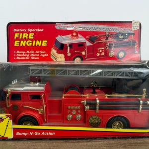 May include: A battery-operated red toy fire engine with a silver ladder, housed in a rectangular box. The packaging features the text Battery Operated FIRE ENGINE, Bump-N-Go Action, Flashing Dome Light, and Realistic Siren in white and yellow.