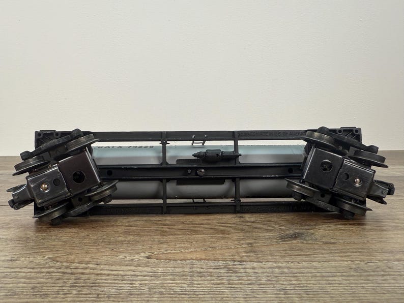 K&ouml;nnte beinhalten: Ein schwarz-grauer Modellzugwagen mit zwei Rads&auml;tzen. Der Wagen hat einen zylindrischen Tank und einen schwarzen Rahmen. Das Modell ist vor einem hellen Hintergrund platziert.