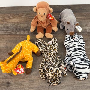 May include: A collection of plush animal toys, including a brown monkey, a gray rhino, a yellow giraffe, a leopard-print cat, and a black and white zebra. Each toy has a tag attached.