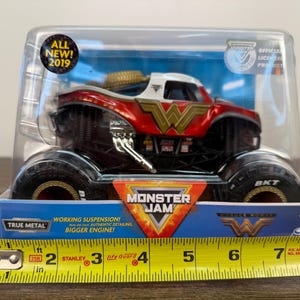 May include: A Wonder Woman Monster Jam truck toy in a clear plastic package. The truck is red, white, and gold with the Wonder Woman logo. The package includes the text "ALL NEW! 2019" and "MONSTER JAM". The truck has large black tyres.