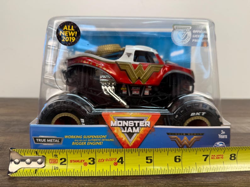 May include: A Wonder Woman Monster Jam truck toy in a clear plastic package. The truck is red, white, and gold with the Wonder Woman logo. The package includes the text "ALL NEW! 2019" and "MONSTER JAM". The truck has large black tyres.