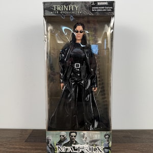 May include: A Trinity action figure from the Matrix movie, packaged in a clear plastic box. The figure is dressed in a long, black coat with a belt and sunglasses. The box has the word "TRINITY" at the top and "MATRIX" at the bottom.