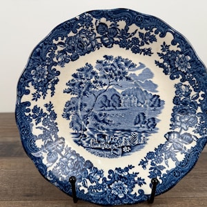 May include: A blue and white decorative plate with a landscape scene in the center. The plate has a scalloped edge and floral border. The landscape scene depicts trees, a building, and a body of water.