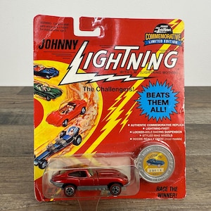 May include: A red Johnny Lightning die-cast toy car in its original packaging. The packaging is red with the words "Johnny Lightning" and "The Challengers" printed on it. The packaging also includes the text "Beats Them All!"