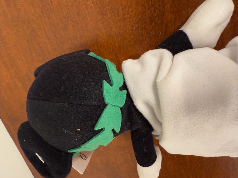 May include: Black and white plush toy with a green felt hat and a white felt body. The toy has a tag that says "Made in Korea".