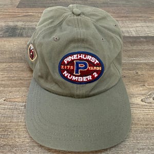 May include: Olive green baseball cap with a curved brim. The cap features an embroidered patch with the text "PINEHURST 7,175 YARDS NUMBER 2" in red, white, and blue. A smaller patch is on the side.