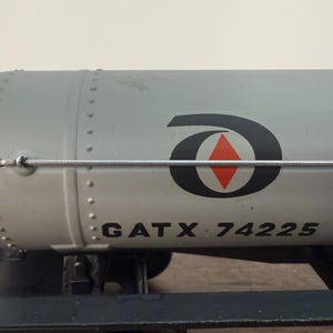 K&ouml;nnte beinhalten: Ein grauer Modell-Eisenbahnkesselwagen mit dem Text "GATX 74225" auf der Seite. Der Kesselwagen hat ein schwarz-rotes Logo und ist auf einem schwarzen Fahrgestell montiert. Der Kesselwagen ist ein Modell eines Eisenbahnwagens.