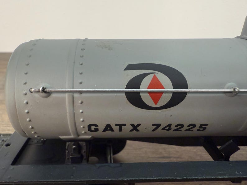 K&ouml;nnte beinhalten: Ein grauer Modell-Eisenbahnkesselwagen mit dem Text "GATX 74225" auf der Seite. Der Kesselwagen hat ein schwarz-rotes Logo und ist auf einem schwarzen Fahrgestell montiert. Der Kesselwagen ist ein Modell eines Eisenbahnwagens.