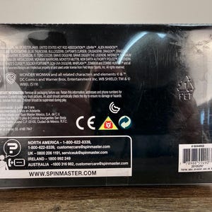May include: A black box with text and logos, including "Wonder Woman" and "DC Comics." The box has a barcode and contact information for Spin Master. The box is made of a clear plastic.