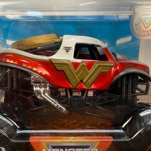 May include: A red, white, and gold Wonder Woman monster truck toy. The truck features the Wonder Woman logo and name, a gold spare tyre, and black wheels. The toy is in its original packaging.