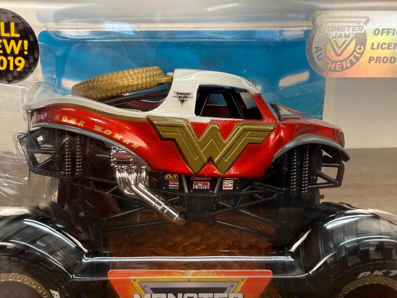 May include: A red, white, and gold Wonder Woman monster truck toy. The truck features the Wonder Woman logo and name, a gold spare tyre, and black wheels. The toy is in its original packaging.