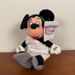 May include: A white plush Mickey Mouse wearing a white robe with a green bow tie. The plush toy is holding a tag that says "The Disney Store".