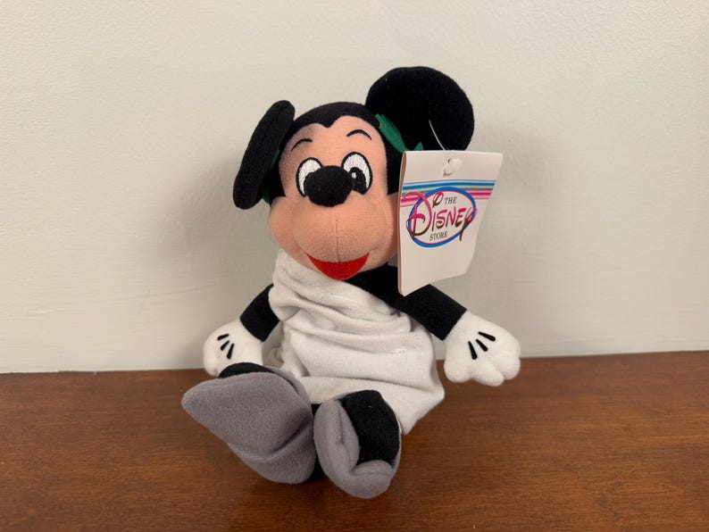 May include: A white plush Mickey Mouse wearing a white robe with a green bow tie. The plush toy is holding a tag that says "The Disney Store".