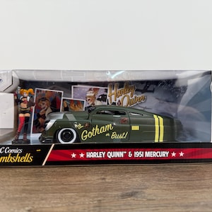 May include: A die-cast model of a 1951 Mercury car, olive green with yellow stripes, in a clear display box. The car has the text "Gotham or Bust!" and is part of the DC Comics Bombshells series, featuring Harley Quinn.
