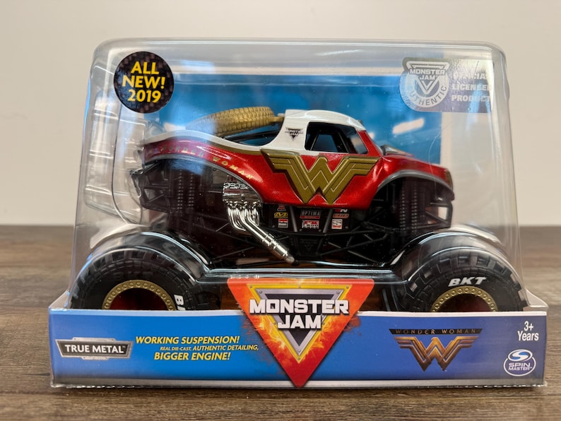 May include: A red, white, and gold Wonder Woman Monster Jam truck toy in a clear plastic package. The toy truck has large black tyres and the Wonder Woman logo. The package includes the text "ALL NEW! 2019" and "MONSTER JAM".