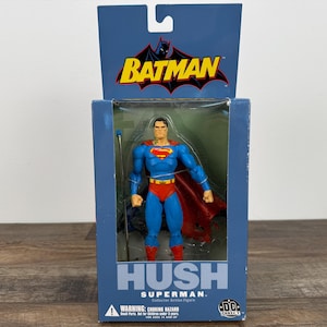 May include: Superman action figure in a blue box with the Batman logo. The figure is in a classic Superman costume with a red cape. The box reads "HUSH SUPERMAN" and includes a warning about choking hazards.