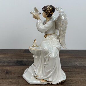 May include: A white porcelain angel figurine with gold accents. The angel is holding a dove and has large, detailed wings. The figurine is seated and has a serene expression. The angel's hair is adorned with a gold headpiece.