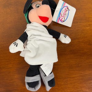 May include: A plush Mickey Mouse toy dressed in a white toga with a green laurel wreath on his head. The toy is wearing sandals and has a red mouth. The toy has a tag that says "The Disney Store".