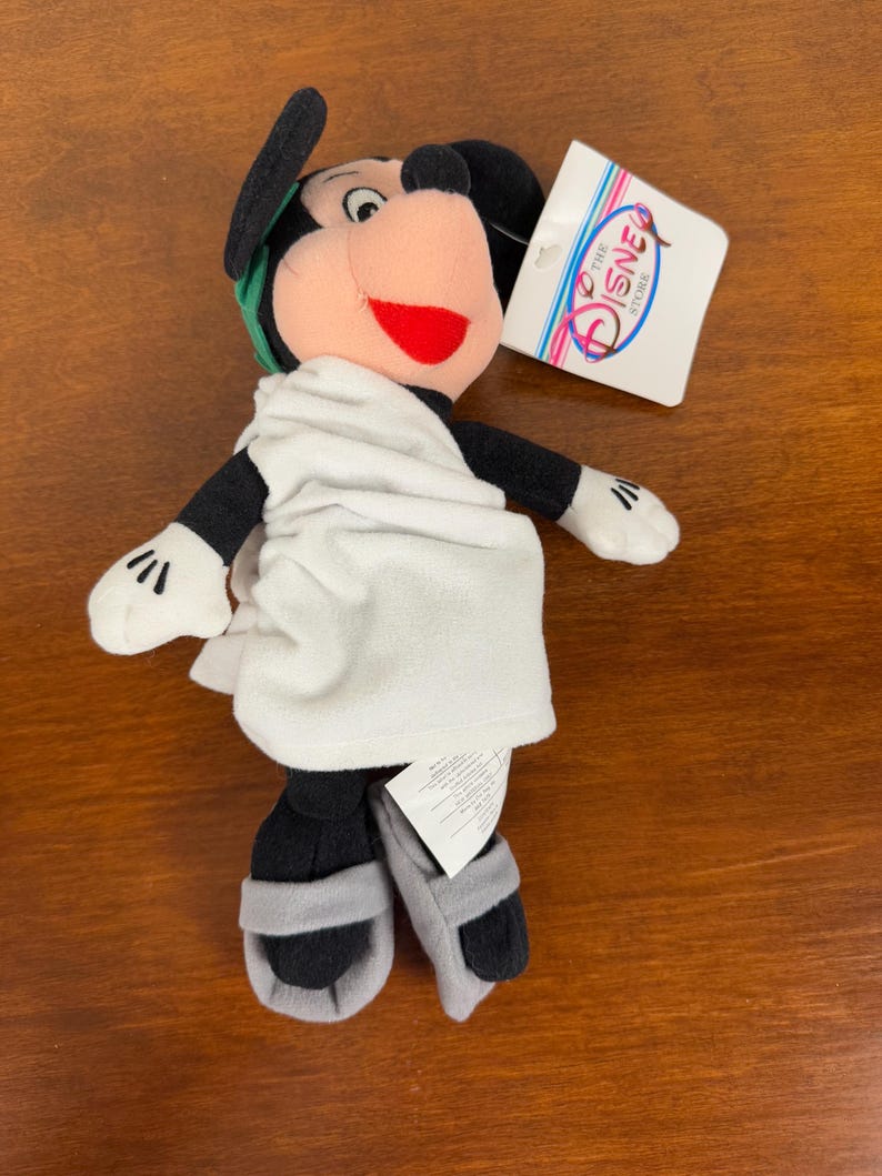 May include: A plush Mickey Mouse toy dressed in a white toga with a green laurel wreath on his head. The toy is wearing sandals and has a red mouth. The toy has a tag that says "The Disney Store".