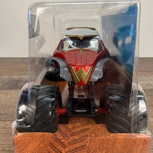 May include: A red, gold, and grey monster truck toy with large black tyres. The truck is in a clear plastic package. The front of the truck has a gold "W" emblem. The packaging sits on a brown surface.