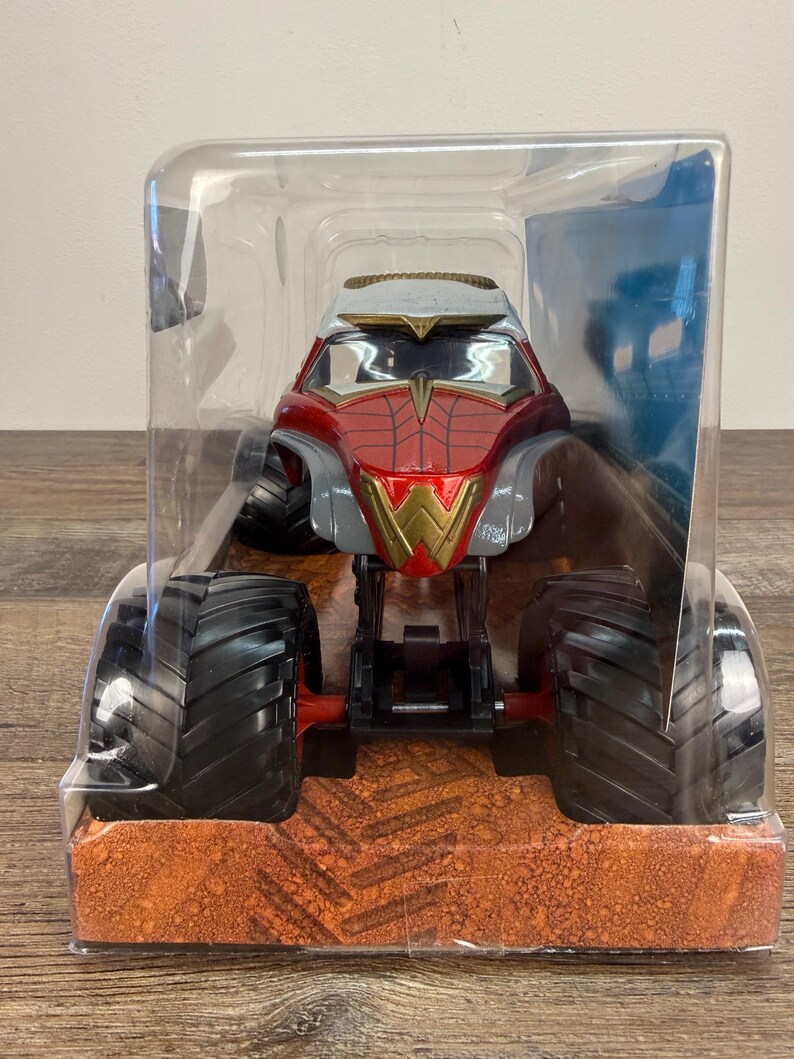 May include: A red, gold, and grey monster truck toy with large black tyres. The truck is in a clear plastic package. The front of the truck has a gold "W" emblem. The packaging sits on a brown surface.