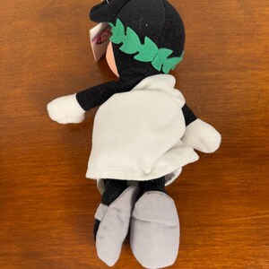 May include: Black and white plush toy with a green laurel wreath on its head. The toy is wearing a white robe and grey sandals.