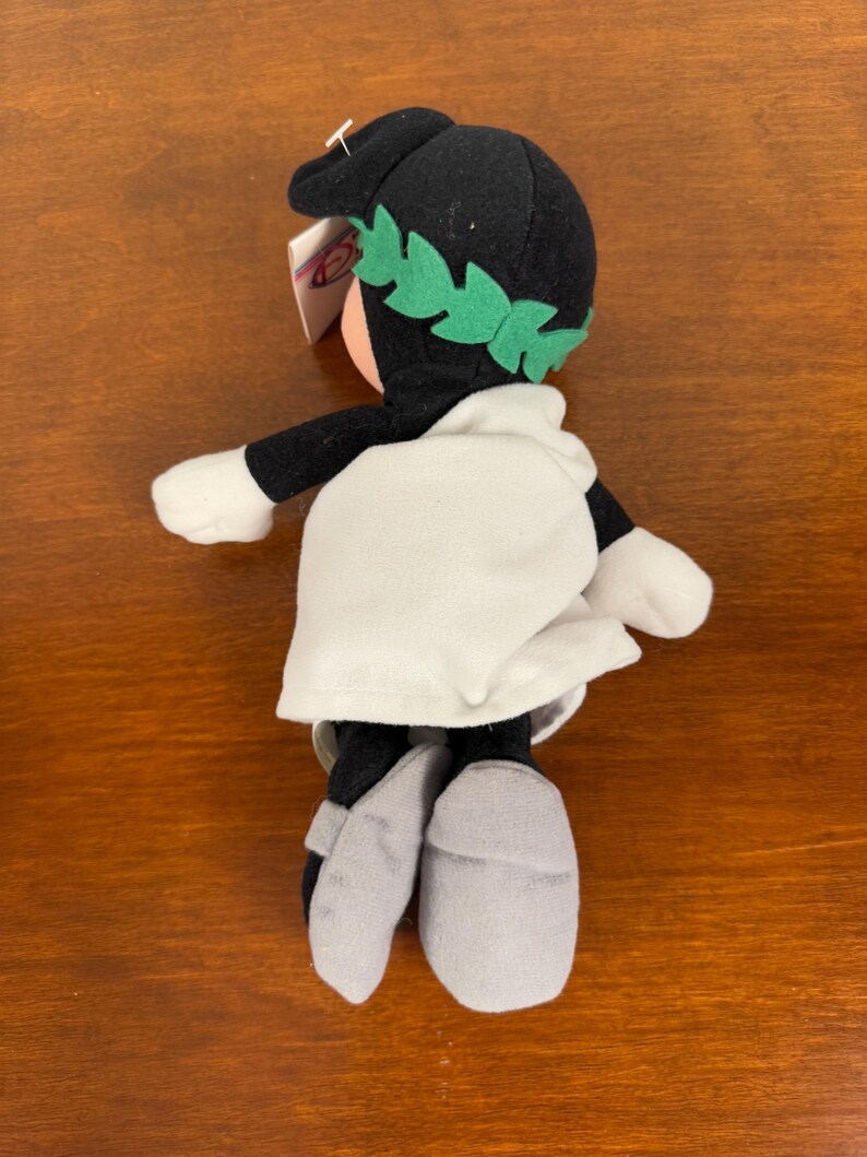 May include: Black and white plush toy with a green laurel wreath on its head. The toy is wearing a white robe and grey sandals.