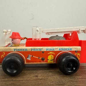 May include: A vintage Fisher-Price fire engine toy featuring a red plastic body, a white extendable ladder, and large black wheels. The side of the truck displays the text FISHER PRICE FIRE ENGINE, along with illustrations of a silver axe and a yellow fire extinguisher.