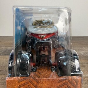 May include: A toy monster truck in a clear plastic package. The truck has large black tyres, a red and silver cab, and a detailed engine. The packaging sits on a brown surface.