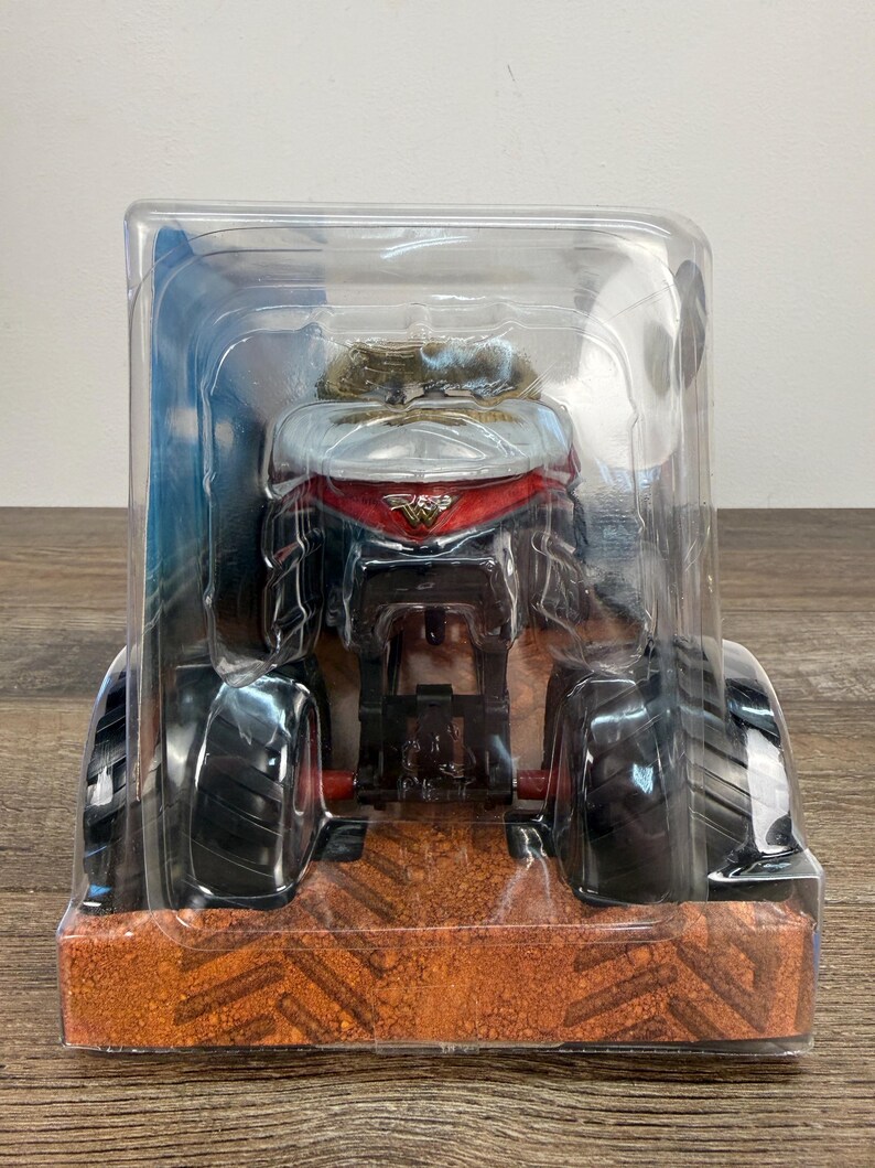 May include: A toy monster truck in a clear plastic package. The truck has large black tyres, a red and silver cab, and a detailed engine. The packaging sits on a brown surface.