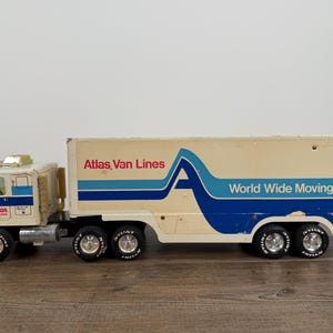 May include: Vintage toy truck with the words "Atlas Van Lines" and "World Wide Moving" printed on the side. The truck is white and beige with blue stripes and accents. The truck has six wheels.