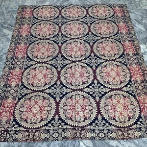 Op de afbeelding: Een groot, rechthoekig textiel met een ingewikkeld patroon. Het ontwerp bevat herhaalde cirkelvormige motieven in tinten bordeauxrood, crème en marineblauw. De randen zijn afgezet met bloemen en geometrische patronen, wat een gedetailleerde en sierlijke uitstraling creëert.
