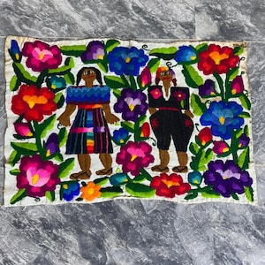 May include: Embroidered textile featuring two figures surrounded by colorful flowers. The figures are dressed in traditional clothing, with vibrant patterns and colors. The flowers include shades of red, pink, blue, and purple, set against a white background.